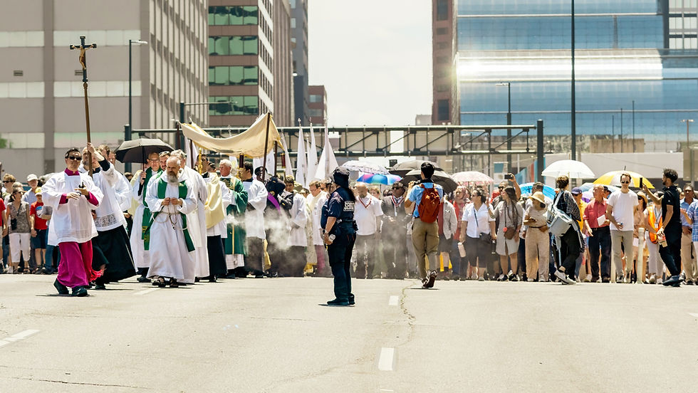 Procession on a city street with clergy in robes, one holding a cross. Crowd follows under colorful umbrellas. Security guards present.