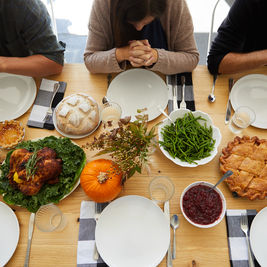 Three people with hands clasped in prayer around a table set with Thanksgiving foods like turkey, pie, and salad. Warm, inviting atmosphere.