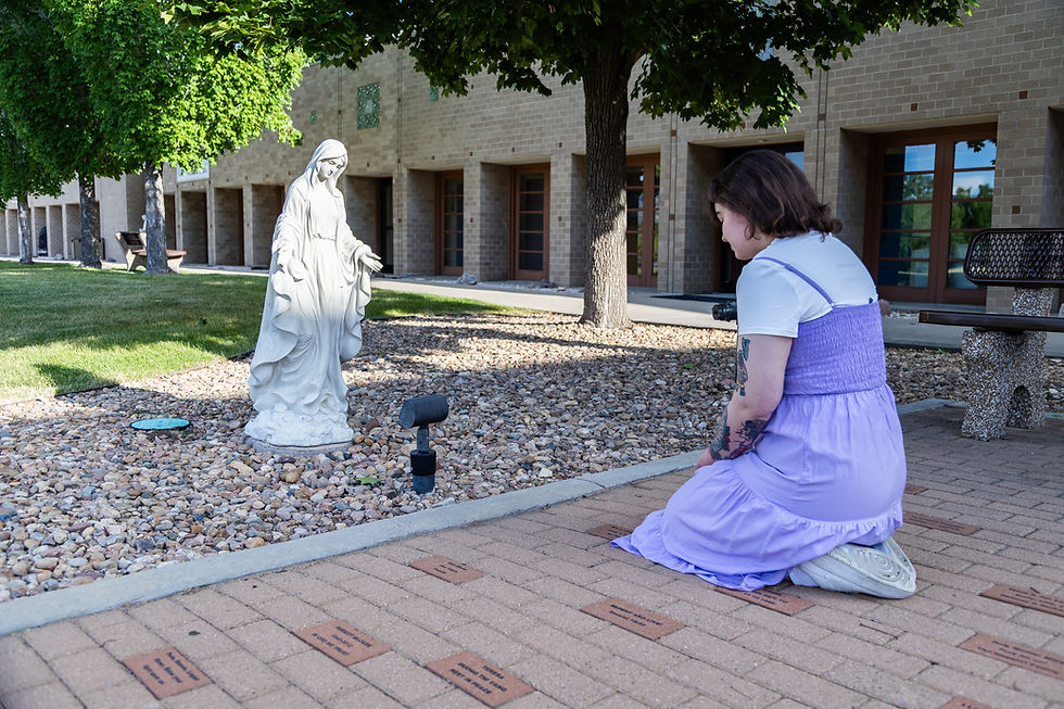 A young woman kneels in prayer on paving stones in front of a white stone statue of Mary, the Mother of God. Patio stones, greenery and trees are seen in the background. The mood is peaceful.