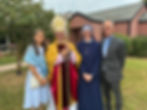 Four people smiling outside a brick building: a woman in blue, a bishop in gold and red attire, a nun in navy, and a man in a suit.