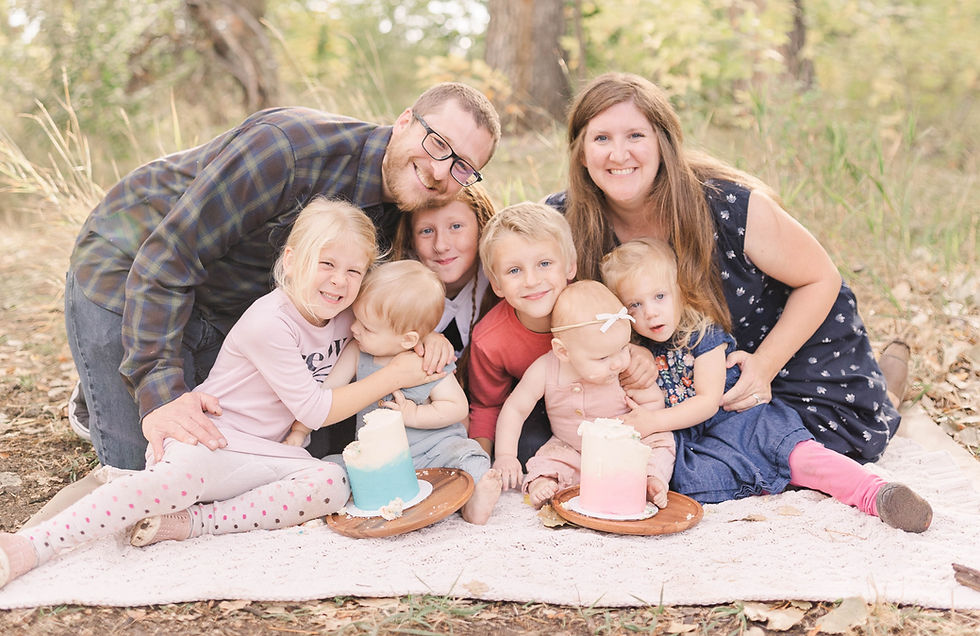 A family of seven smiling in a grassy setting. Two cakes with blue and pink frosting sit on plates. Warm, joyful atmosphere.
