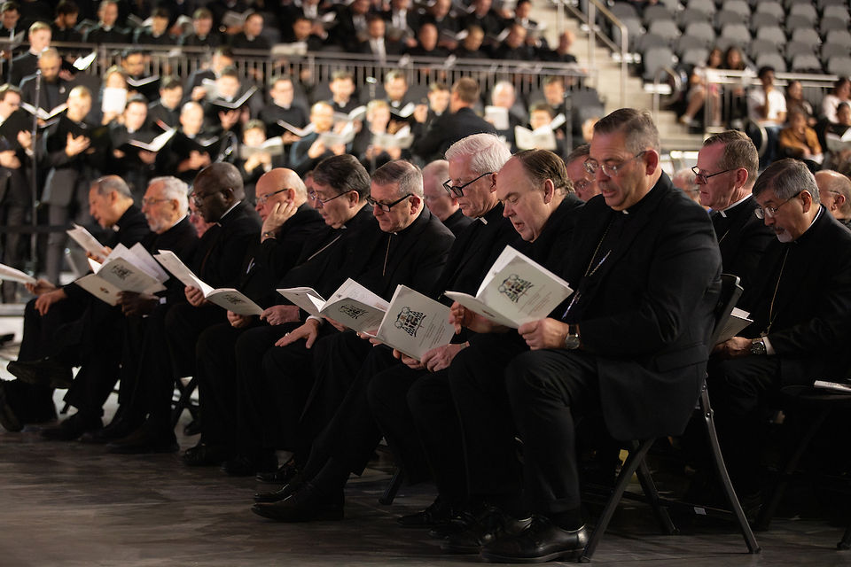 Group of men in black clerical attire seated, reading from booklets. Large choir in background, concert hall setting. Formal atmosphere.