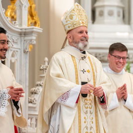 Three clergymen in ornate robes stand in a church, smiling. The central figure wears a detailed mitre. Decorative white and gold background.
