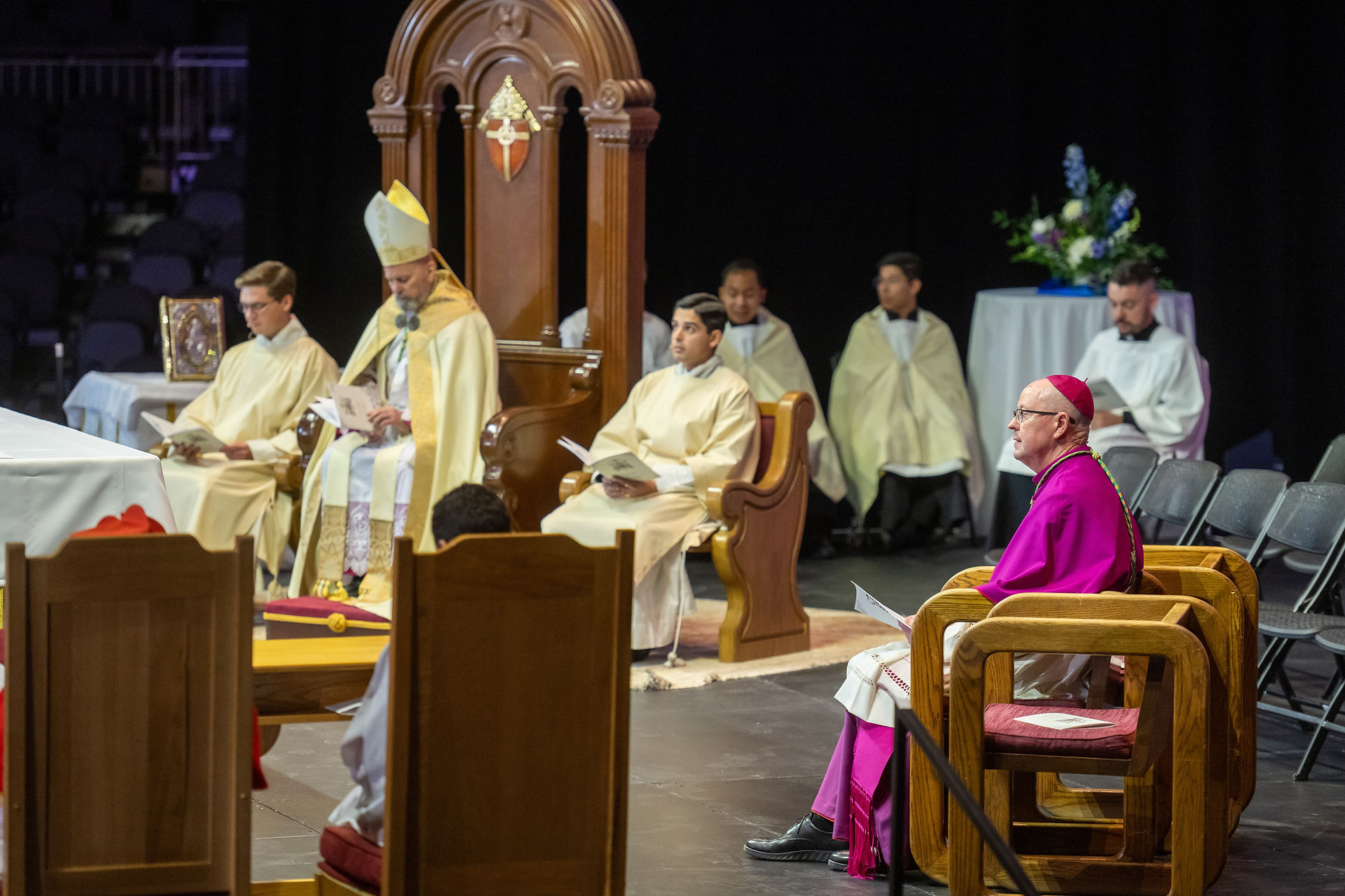 Clergy in cream vestments sit in chairs on a stage during a liturgy, while a bishop in magenta sits nearby holding a program.