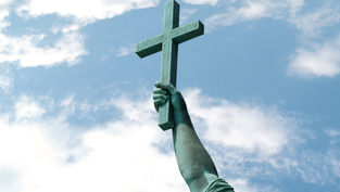 Bronze statue arm holding a cross aloft against a blue sky with scattered clouds