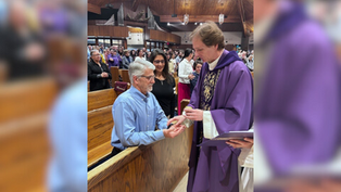 Priest in purple robe blesses the hands of a man in blue at a church. Congregation stands in pews. Wooden ceiling and organ pipes visible.