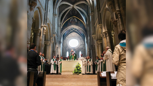 Clergy conduct a service in a grand cathedral with high arches, wearing white and green robes. Attendees watch solemnly in the foreground.