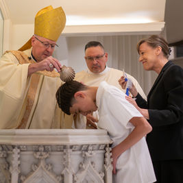 Bishop pours water over a bowed boy during a baptism, while a smiling woman supports him in a church.