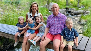 Smiling adults and children sit on a wooden bridge over a stream in a lush, green park. Bright and cheerful atmosphere.