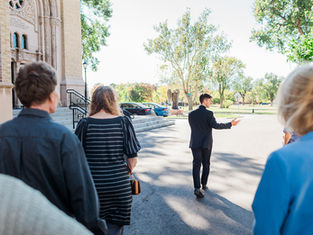 A group follows a man in a suit pointing ahead, near a church with ornate architecture. Trees and parked cars in the sunny background.