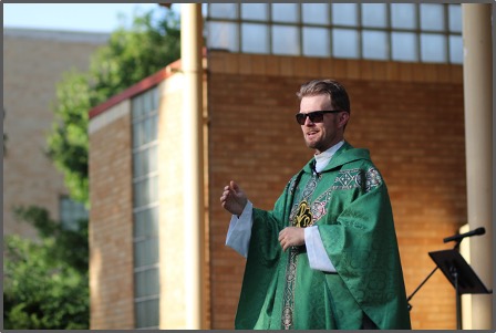 A person in green vestments speaks outdoors, with trees and a building in the background. Sunglasses add a casual touch to the solemn setting.