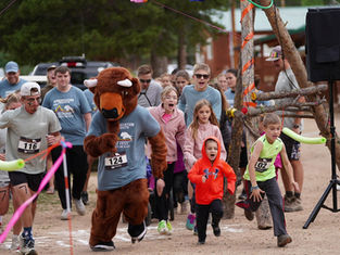 Runners, including a person in a buffalo costume, race outdoors on a dirt path, surrounded by spectators. Energetic atmosphere, colorful attire.