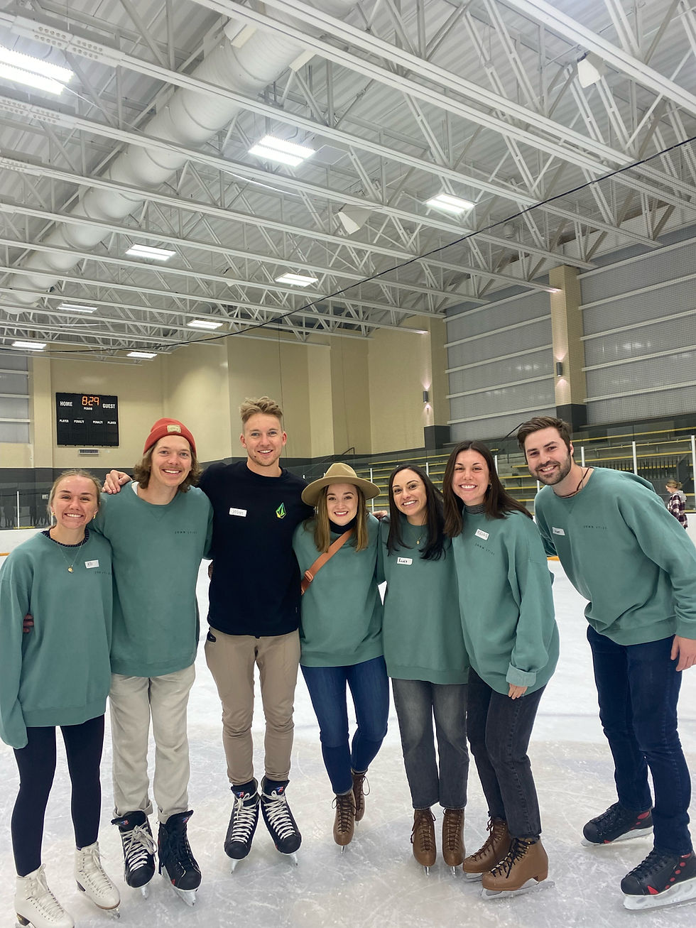 Seven smiling people in casual clothes and skates pose on an indoor ice rink, with a high ceiling and visible scoreboard in the background.