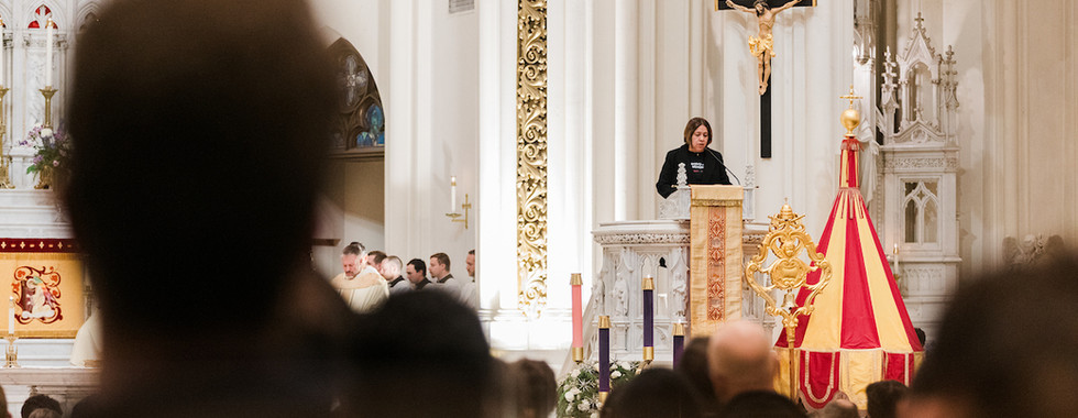 A woman speaks at a pulpit inside a grand church with ornate pillars. People sit in pews, listening. A crucifix and decorative red and gold element are visible.