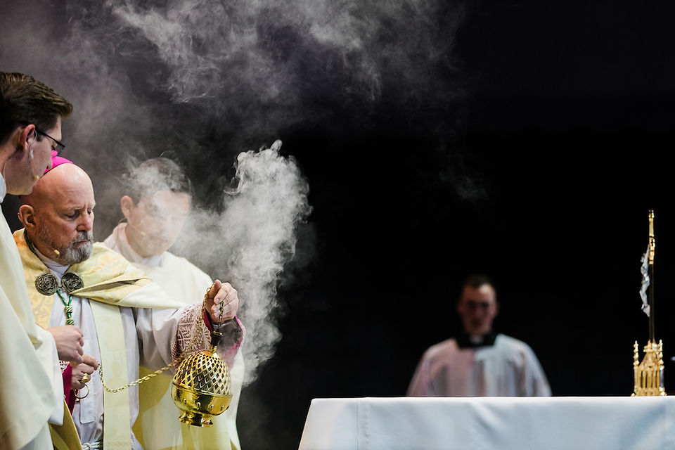 Clergy in ceremonial robes swing a golden censer, releasing smoke. Dark background, altar with a white cloth. Solemn mood.