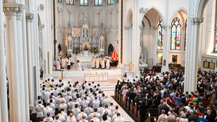 Clergy in white robes lead a ceremony in a grand cathedral with stained glass windows. A large congregation watches solemnly.