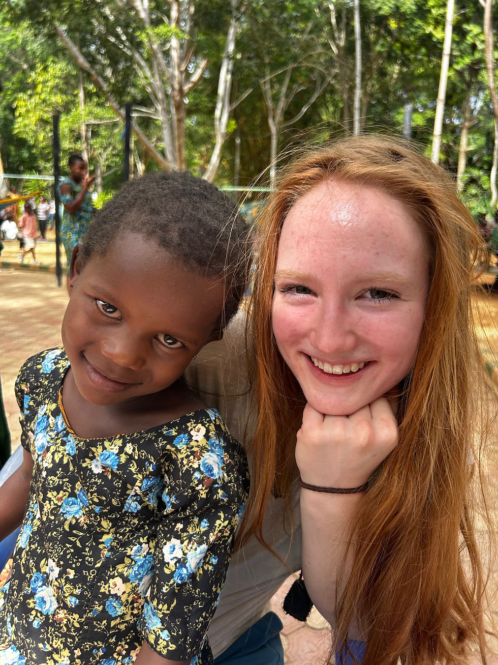Two girls smiling outdoors. The girl on the left wears a floral dress, while the one on the right has red hair. Trees in the background.