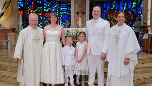 A family and two clergy in white attire smile inside a church with colorful stained-glass windows. The mood is joyful and celebratory.