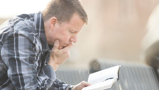 A man in a plaid shirt reads thoughtfully outdoors on a sunny day, holding a book with focus and concentration.