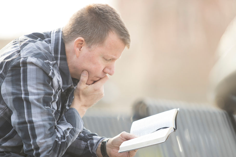 A man in a plaid shirt reads thoughtfully outdoors on a sunny day, holding a book with focus and concentration.