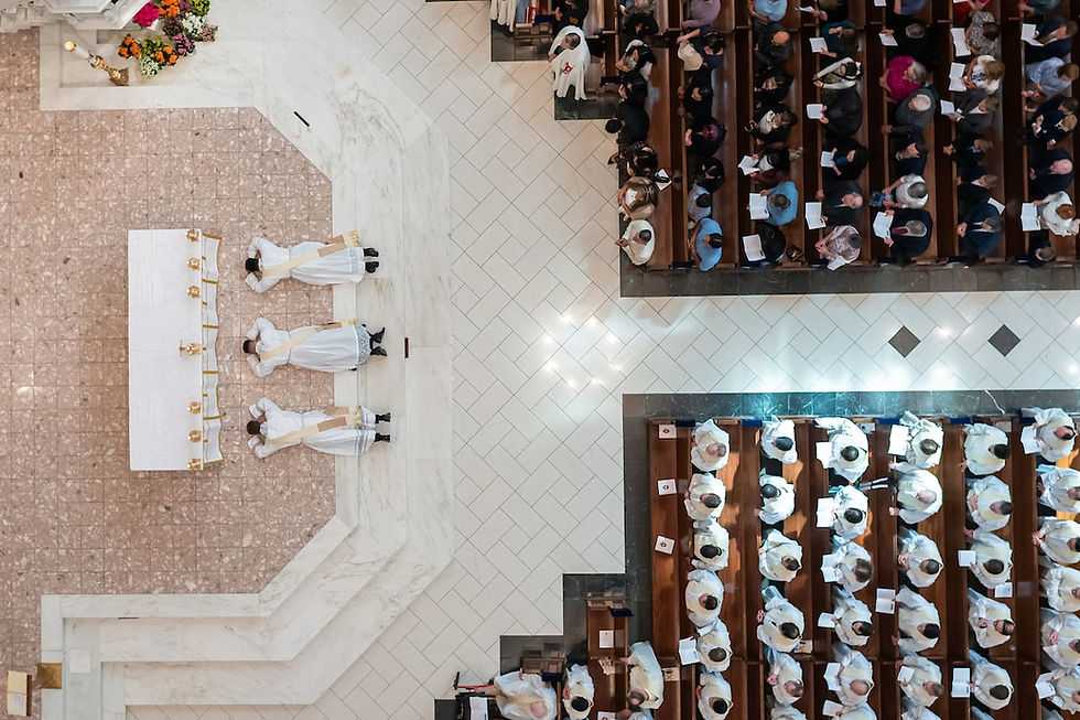 Aerial view of three priests lying prostrate before an altar in a church, surrounded by seated congregation, highlighting ceremonial solemnity.