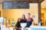 Three smiling baristas behind a coffee shop counter with a menu board above. Display of chips, a payment terminal, and a minimalist decor.