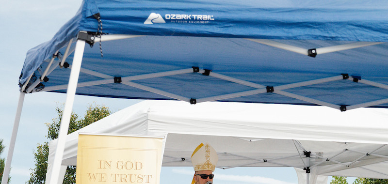 Bishop speaks under a blue canopy to a seated audience. Yellow banner reads "IN GOD WE TRUST." Outdoor setting with trees in the background.