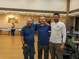 Three smiling men in casual clothes pose in a hall with wooden floors. Tables in the background have food trays. Warm lighting and a relaxed mood.