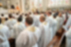 Priests in white robes with cream collars standing in a church. Stained glass windows and altar visible. Formal, solemn atmosphere.