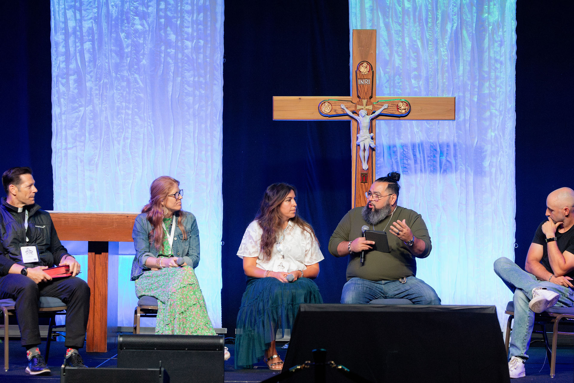 Panel of five people speaking on a church stage before a large crucifix and blue-lit curtains, with attentive, serious expressions