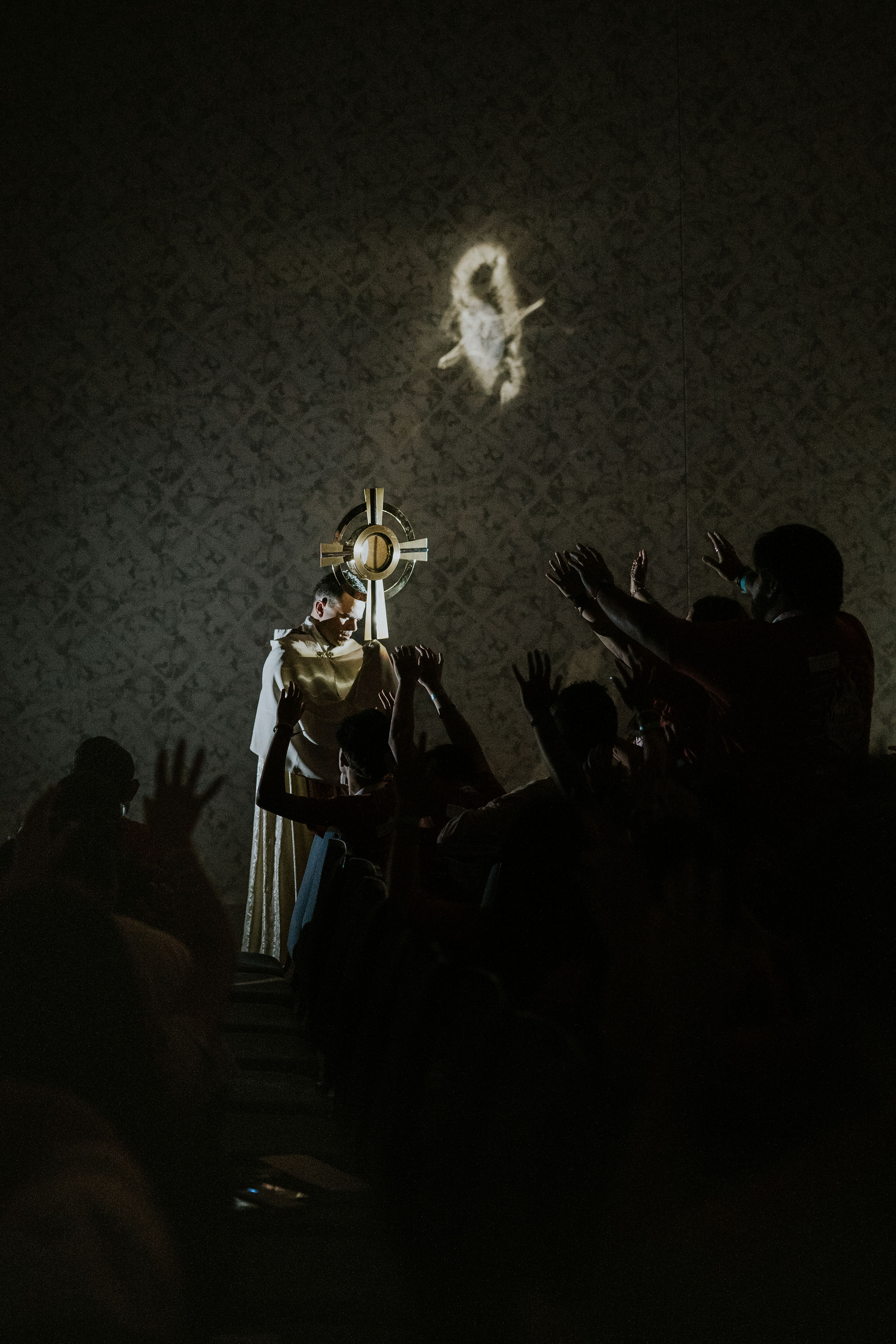 A person in white robes holds a large gold cross in a dim room. Hands reach towards the light, casting dramatic shadows on the wall.