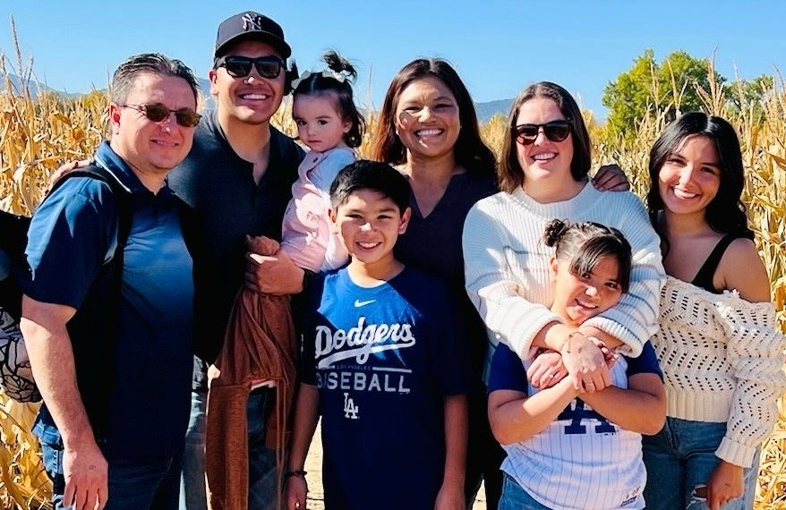 A group of smiling people, including kids, pose outdoors in a cornfield. A boy wears a blue Dodgers T-shirt. Bright, sunny day, clear sky.