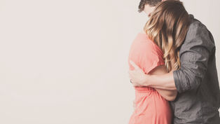 Couple embraces warmly, both in casual attire; the woman in pink, man in grey. Light background, soft and intimate mood.