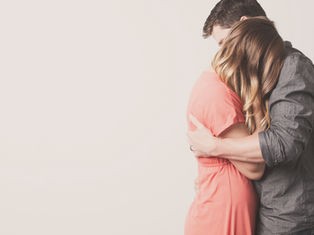 Couple embraces warmly, both in casual attire; the woman in pink, man in grey. Light background, soft and intimate mood.