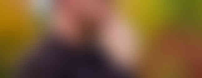 A man in a black jacket and gray cap with "N" thoughtfully observes. Autumn trees and a building blur in the background.