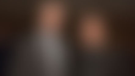 Two men in suits smile at a formal event. The background has patterned artwork, and a sign with text is partially visible. Atmosphere is friendly.