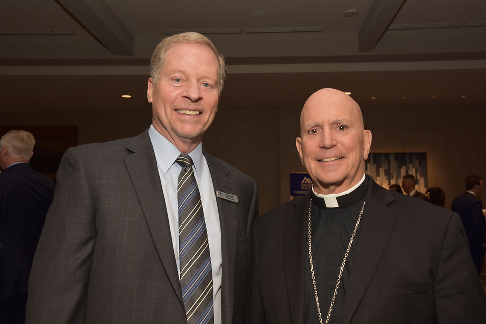 Two men in suits smile at a formal event. The background has patterned artwork, and a sign with text is partially visible. Atmosphere is friendly.