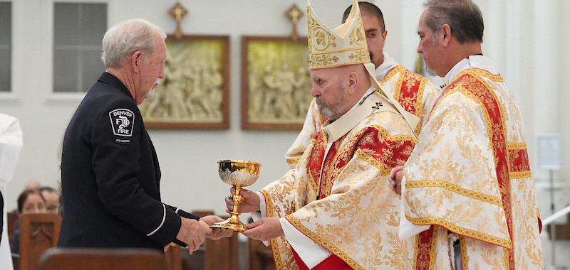 A bishop in ornate vestments receives a chalice from a man in a uniform inside a cathedral with stained glass windows. The mood is solemn.