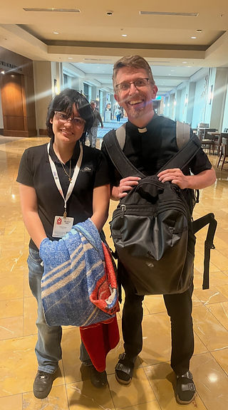 Two people stand smiling in a hallway. One holds a striped blanket, and both wear name badges. The setting is warmly lit with a tiled floor.