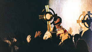 Catholic priest holds a golden monstrance before raised hands in a dim, glowing worship service.