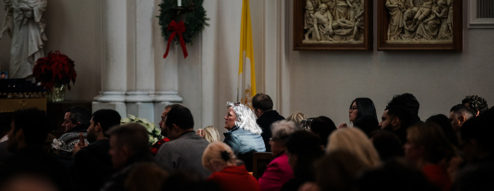 People seated in a church during a service, some with heads bowed. Religious statues and artwork in the background. Somber atmosphere.
