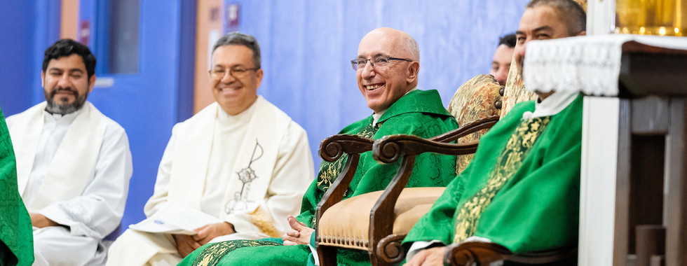 Smiling priests in green and white vestments sit in a church hall against a blue wall during a religious ceremony.