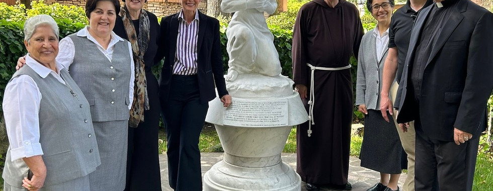 A group of men and women, including clergy and religious sisters, stand together outdoors near a statue during a visit to a church office or courtyard.