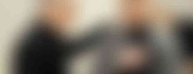 Two men in clerical attire converse warmly, one with a hand on the other's shoulder. Background shows a table with cups and plates.