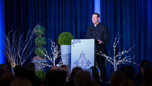 Man in black clerical attire speaks at a lectern with decorative designs. Blue curtains and lit branches set a formal, serene mood.