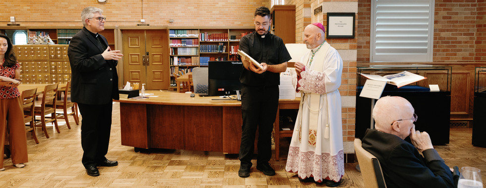 Priest shows a book to a bishop in a library with brick walls. Two others observe, one seated. Shelves and wooden furniture in background.