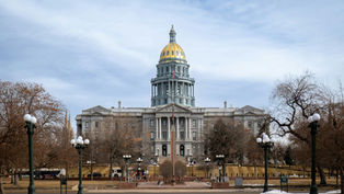 Gray domed capitol building with a golden top against a cloudy sky. Trees and streetlights line the walkway. Sparse visitors in the foreground.