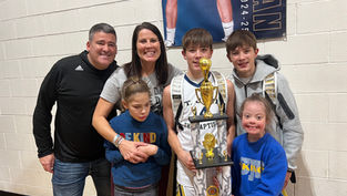 A group of smiling people poses with a sports trophy in a gym. Two individuals wear "BE KIND" shirts. A poster hangs in the background.