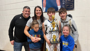 A group of smiling people poses with a sports trophy in a gym. Two individuals wear "BE KIND" shirts. A poster hangs in the background.