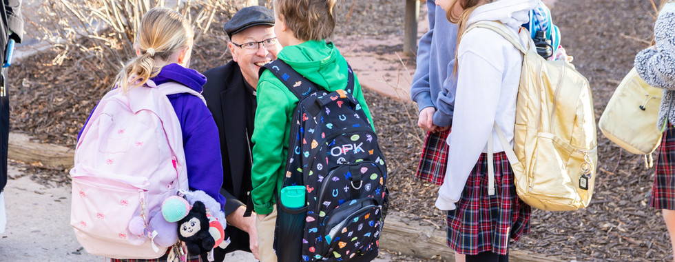 Man kneeling and smiling at four children with colorful backpacks, outdoors. Kids wear school uniforms with plaid skirts. Autumn setting.